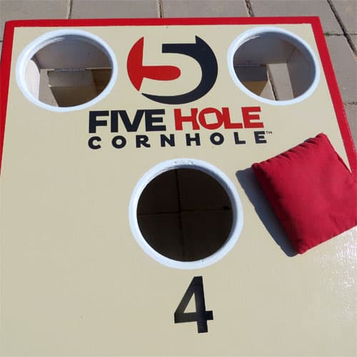 Top of a 5-Hole cornhole board showing two corner holes, a numbered hole, and a red bag