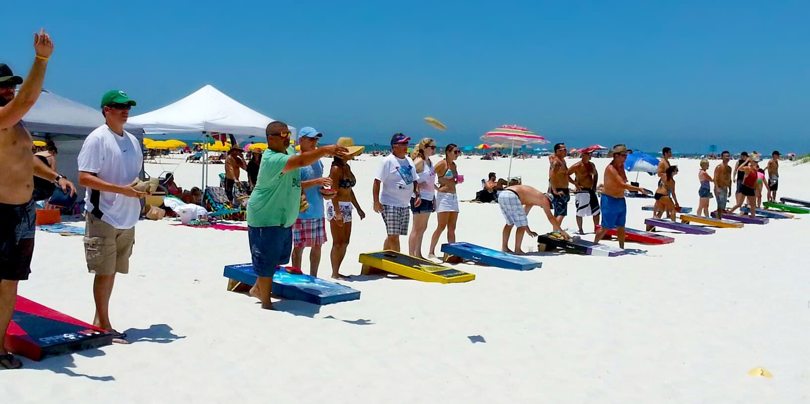 Players tossing bags at a Custom Corntoss beach cornhole tournament with rows of boards