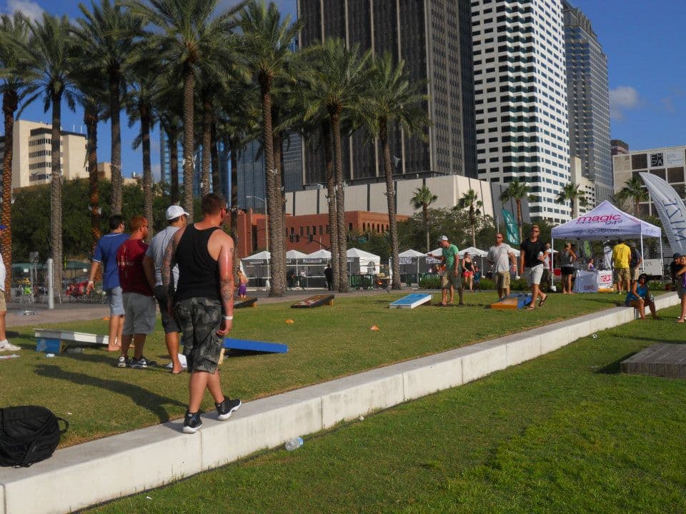 Players at an outdoor cornhole tournament in a downtown park with a sponsor tent