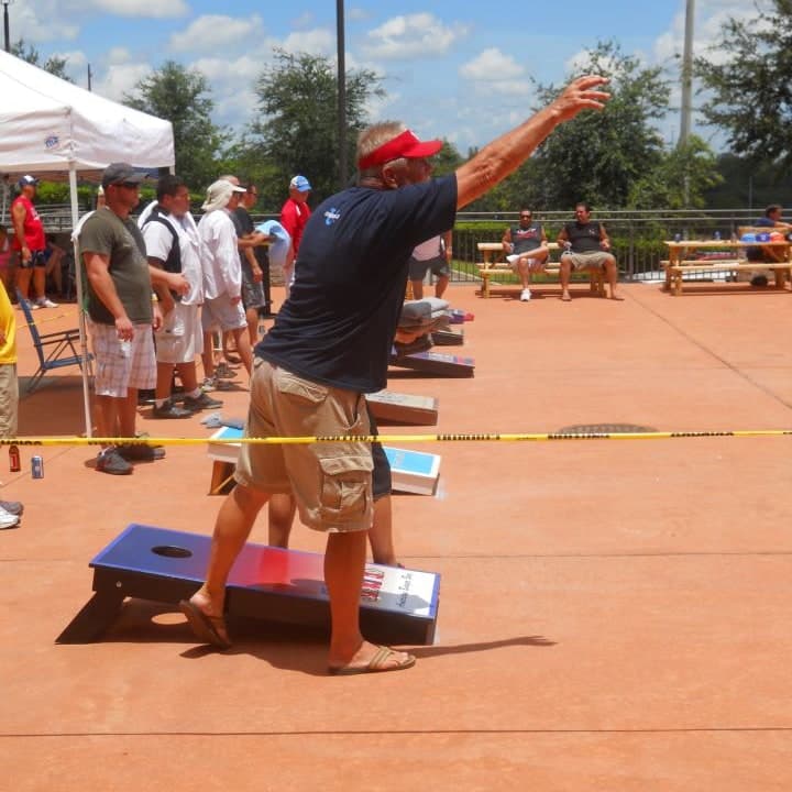Man in a red visor tossing a cornhole bag underhand at an outdoor cornhole tournament